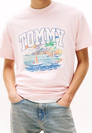 Person iført en lyserød T-shirt med farverig grafik af en båd, palme og ø samt teksten "TOMMY Jeans".