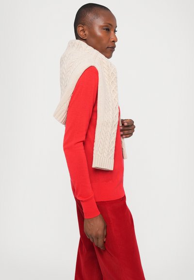 Red long-sleeve sweater paired with red corduroy pants, complemented by a light beige knitted scarf draped over the shoulders, featuring a textured pattern.