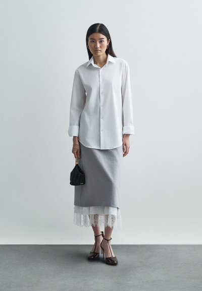 Bijela košulja s dugim rukavima, siva olovka suknja s čipkastim rubom, crna torba i smeđe cipele s visokom petom. Model stoji protiv neutralne pozadine.