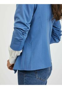 Modrý oblekový kabát se zřasenými rukávy, vrstvený přes bílou texturovanou blůzu. Zadní pohled ukazuje štíhlý design a přizpůsobenou siluetu.