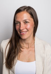 Ritratto di una donna con lunghi capelli castani, che indossa una maglietta bianca e un blazer beige chiaro, messo in risalto da una collana con catena dorata. Sfondo neutro.