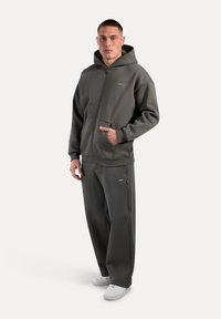 Grå hætte-trøje med lynlås og lomme foran, parret med matchende joggers. Fremstillet i blødt stof med en afslappet pasform. Hvide sneakers.