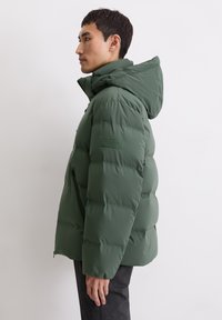 Doudoune verte avec capuche, présentant un design matelassé, des poches zippées et un patch logo discret sur la manche. Fini doux et mat.