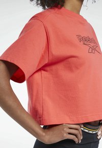 Korálové zkrácené tričko vyrobené z bavlny s krátkými rukávy, obsahující černě vyšívané logo Reebok na přední straně a uvolněný střih.