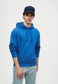 Μπλε φούτερ με κουκούλα και κορδόνι, με ριμπ μανσέτες, που διαθέτει ένα διακριτικό λογότυπο στο στήθος. Συνδυάζεται με ανοιχτό μπλε τζιν και ναυτικό καπέλο.