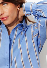 Chemise bleue à boutons avec des rayures verticales blanches et beige. Manches longues avec détail noué et col classique. Texture douce.