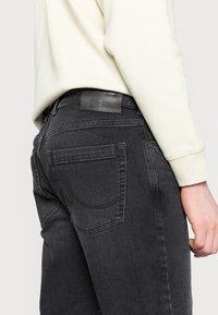 Jeans de mezclilla gris con bolsillos traseros, costuras sutiles y una etiqueta de marca negra en la cinturilla. La tela tiene una textura suave.