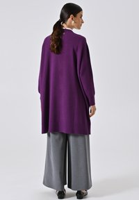 Langer lila Strickcardigan mit lockerer Passform, geripptem Saum und Rückendetails. Kombiniert mit weiten grauen Hosen und schwarzen Schuhen.