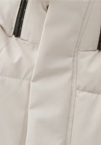 Veste rembourrée beige avec coutures visibles matelassées et une fermeture éclair noire sur le côté gauche.