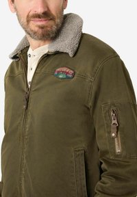 Olivgrüne Jacke mit einem grauen Lammfellkragen, Reißverschluss vorne und einem Patch auf der Brust; mit einer Reißverschlusstasche am linken Ärmel.