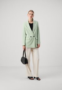 Blazer de doble botonadura verde claro con botones negros, combinado con pantalones de pierna ancha crema y tacones negros. Sosteniendo un bolso negro.
