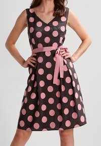 Vestido negro con grandes lunares rosas, escote en V y un lazo rosa en la cintura. Hecho de una tela suave con una falda acampanada y diseño sin mangas.