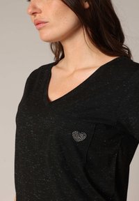 Top noir à col en V avec un tissu texturé, des manches courtes et une poche sur la poitrine gauche ornée d'un cœur en strass.