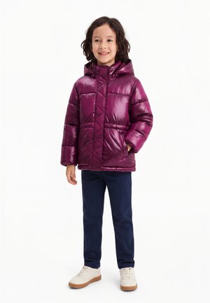 Enfant aux cheveux bouclés portant une doudoune à capuche violet brillant, un pantalon bleu marine et des baskets blanches, debout et souriant.