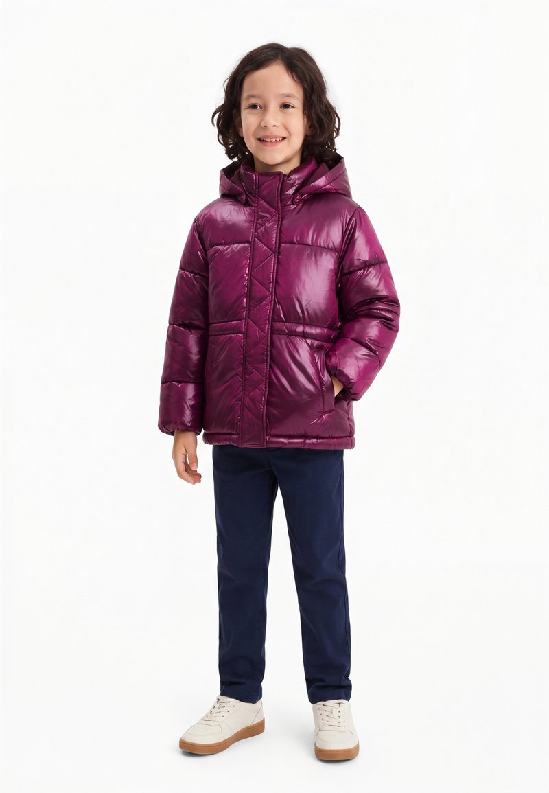 Enfant aux cheveux bouclés portant une doudoune à capuche violet brillant, un pantalon bleu marine et des baskets blanches, debout et souriant.