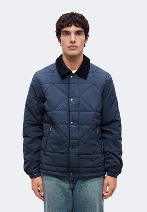 Un homme porte une veste matelassée bleu marine à col noir et un jean bleu, sur un fond clair et uni.