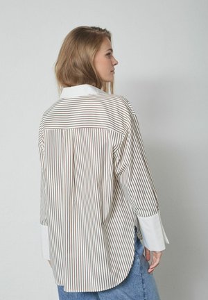 Tânără văzută din profil, purtând o cămașă albă cu dungi verticale maro și manșete albe, asortată cu blugi albaștri, în fața unui perete simplu.