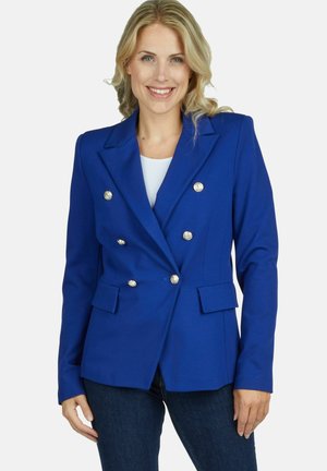Blond vrouw glimlacht, draagt een koningsblauwe dubbelgeknoopte blazer met zilveren knopen en donkerblauwe spijkerbroek, staand tegen een witte achtergrond.