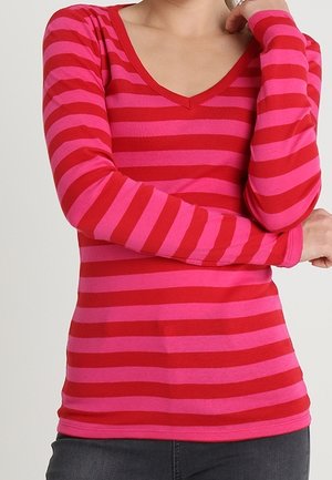 Personne portant une chemise à manches longues rayée horizontalement rouge et rose avec un col en V, bras croisés et pantalon sombre.