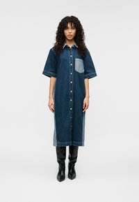 Φόρεμα πουκάμισο από τζιν σε σκούρο μπλε με ανοιχτό μπλε τσέπη στο στήθος, κοντά μανίκια, κλείσιμο με κουμπιά και αντίθετες πλαϊνές λεπτομέρειες. Φορεμένο με μαύρες μπότες μέχρι το γόνατο.