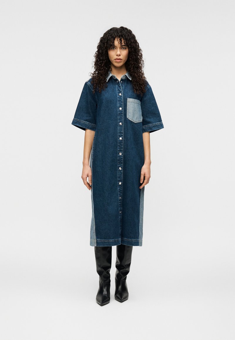 Φόρεμα πουκάμισο από τζιν σε σκούρο μπλε με ανοιχτό μπλε τσέπη στο στήθος, κοντά μανίκια, κλείσιμο με κουμπιά και αντίθετες πλαϊνές λεπτομέρειες. Φορεμένο με μαύρες μπότες μέχρι το γόνατο.