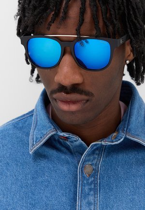 Uomo con dreadlocks che indossa occhiali da sole con montatura nera e lenti specchiate blu e una giacca di jeans blu con colletto abbottonato.