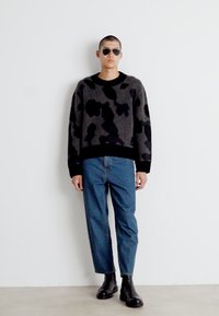 Pulover cu model camuflaj gri și negru, cu guler rotund, asortat cu blugi drepti de un albastru deschis și cizme negre din piele până la gleznă.