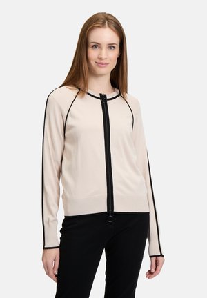 Cardigan beige clair avec des accents noirs et une fermeture éclair à l'avant. Il présente un col rond, des manches longues et une coupe ajustée. Texture douce en tricot.