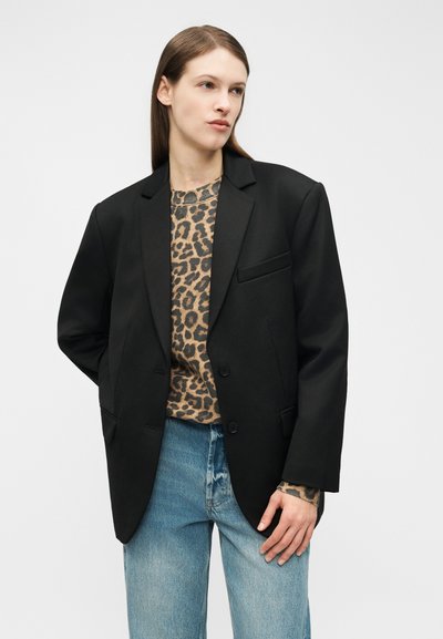 Osoba oblečená v čiernom nadmerne veľkom blejzri, leopardom potlačenom topu a svetlo modrých džínsach, stojaca pred jednoduchým bielym pozadím.