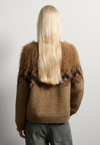 Brauner Strickpullover mit einer flauschigen Textur und schwarzen geometrischen Mustern auf den Schultern. Die langen Haare des Models sind sichtbar.