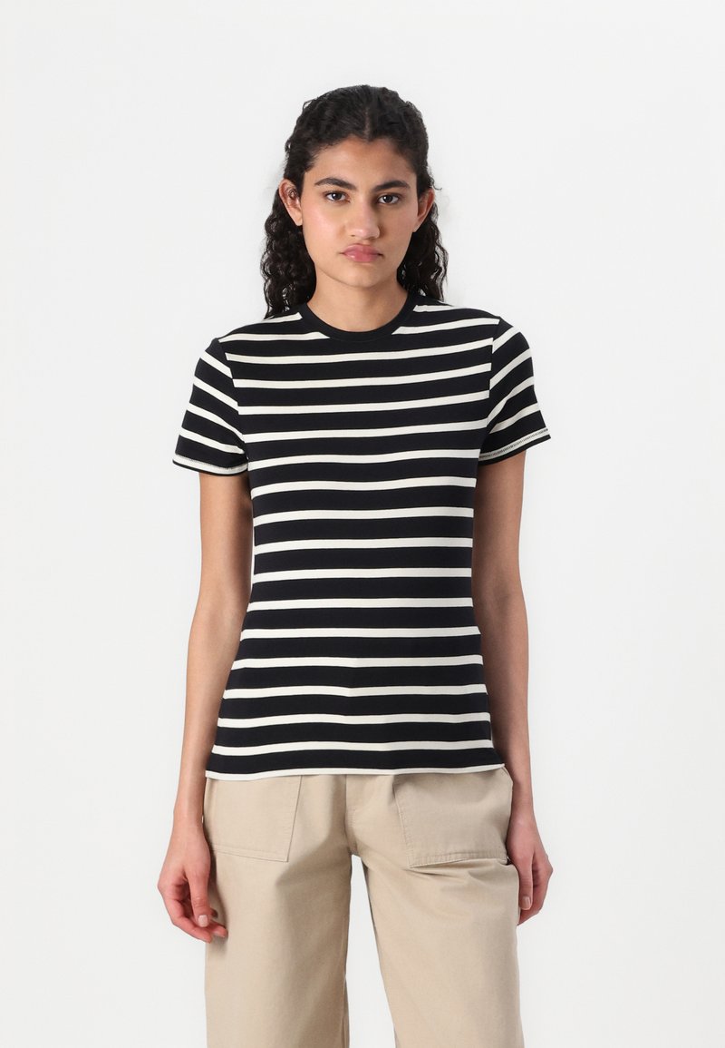 T-shirt a righe bianche e nere con colletto tondo e maniche corte, realizzata in tessuto morbido, abbinata a pantaloni beige chiaro.