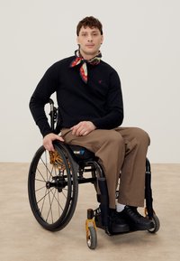 Homme assis dans un fauteuil roulant noir avec des détails orange, portant un pull noir, un pantalon beige, des chaussures noires et un foulard à motifs autour du cou sur un sol neutre.