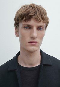 Young man with light brown tousled hair and blue eyes wearing a black shirt and jacket against a plain light background.