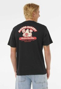Blonde man in een zwart T-shirt met een rood-witte afbeelding van "Board Bakery Rip Curl Surf Shop" en cartoon surfers op de achterkant.