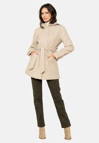 Manteau en mélange de laine beige avec une taille ceinturée, une capuche et des poches latérales, associé à un pantalon vert olive et des bottines beiges.