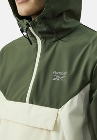 Groene en crèmekleurige Reebok pulloverjack, met een capuchon, een kwart-rits aan de voorkant en een gestructureerde stof met een zichtbaar merklogo.