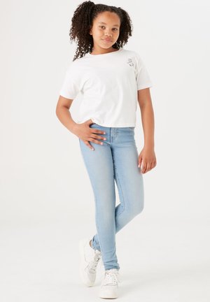 Camiseta blanca cortada con texto, jeans ajustados azules claros y zapatillas blancas con acentos perforados. La modelo está en una pose relajada.