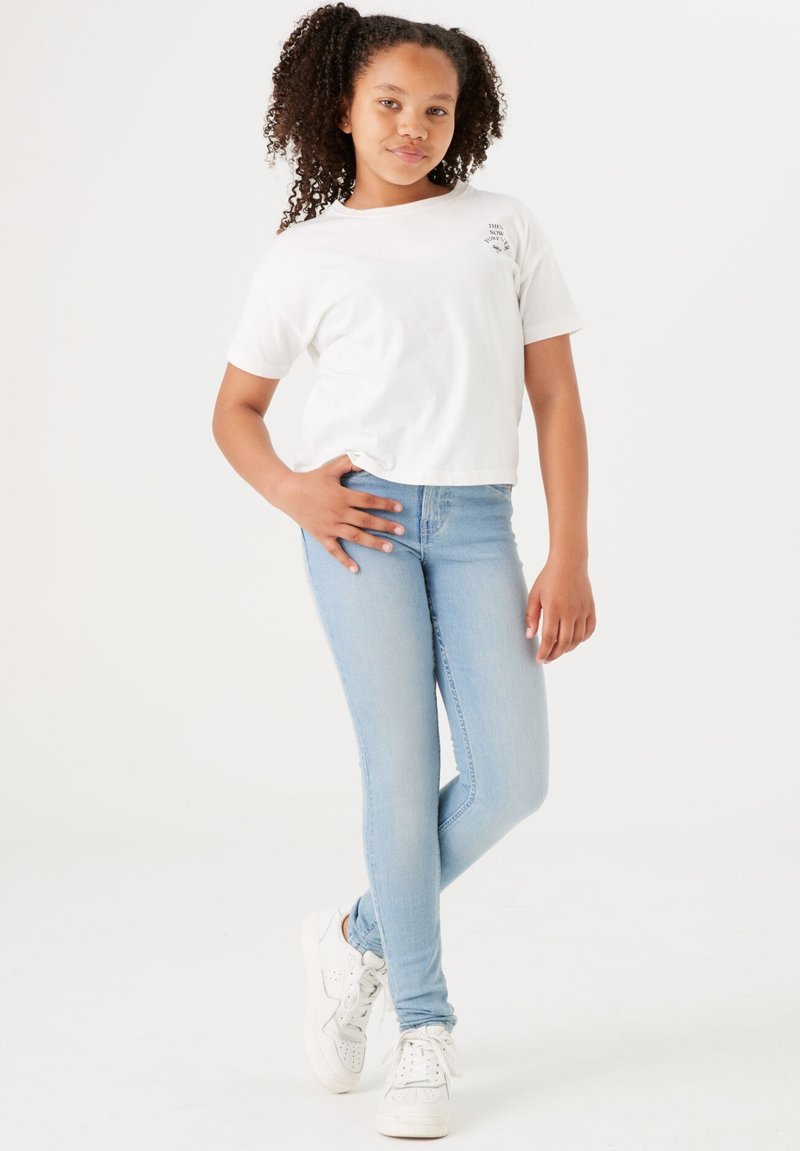Weißes, kurzes T-Shirt mit Schriftzug, hellblaue Skinny-Jeans und weiße Sneaker mit perforierten Akzenten. Model steht in einer entspannten Pose.
