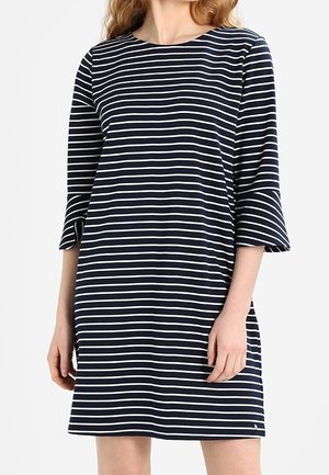 Frau trägt ein marineblau-weiß horizontal gestreiftes Kleid mit dreiviertel Ärmeln und leicht ausgestellten Manschetten.