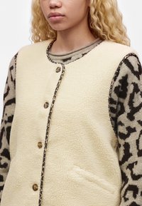 Femme portant une veste sans manches en polaire crème avec fermeture à boutons par-dessus un pull à manches longues imprimé léopard.