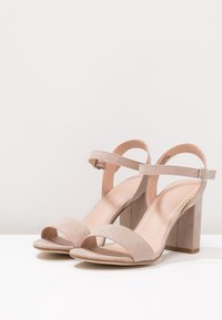 Beżowe zamszowe sandały na obcasie z otwartym noskiem, regulowanym paskiem na kostce oraz kwadratowym słupkiem. Gładka tekstura i minimalistyczny design.