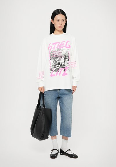 White sweatshirt with pink graphic text and monochrome image, paired with cropped blue denim pants, black handbag, and black shoes.