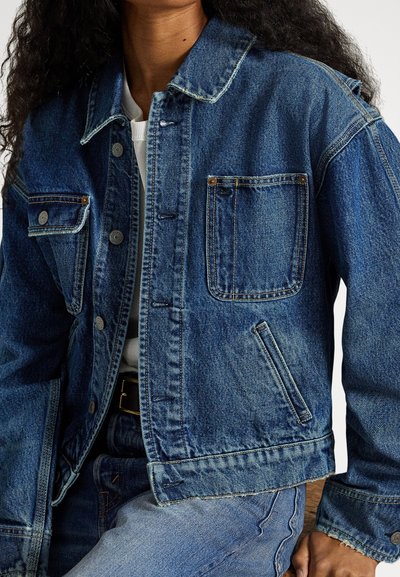 Person som har på seg en blå denimjakke med knappedetaljer, en hvit skjorte og lyseblå jeans, sitter med den ene hånden hvilende på kneet.