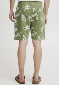 Pantaloni corti verdi con motivo a foglia bianca, cintura elastica e tasca posteriore. Il materiale sembra morbido e con una texture, adatto per un abbigliamento casual.