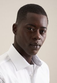 Jeune homme noir aux cheveux courts portant une chemise blanche avec un col à motif, regardant légèrement sur le côté, sur un fond beige.