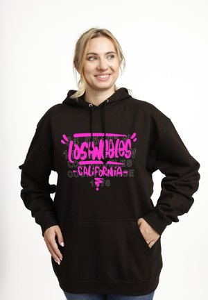 Donna sorridente con capelli biondi che indossa una felpa nera con cappuccio, caratterizzata da un vivace testo in stile graffiti rosa acceso con scritto "Los Angeles California" e numeri sul davanti.