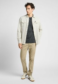 Camisa de botones a rayas beige y blanca sobre una camiseta gris, combinada con pantalones beige claros y zapatillas blancas con rayas negras.