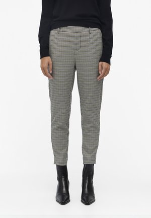 Person iført sort-hvide småternet slim-fit bukser, sort langærmet top og sorte spidse ankelstøvler, stående oprejst.