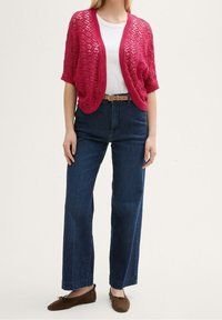Frambozenroze opengewerkte cardigan over een wit T-shirt, gecombineerd met donkerblauwe, wijde, hoge taille jeans en bruine platte schoenen.