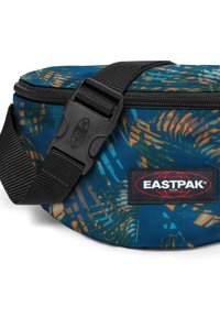 Marsupio blu con motivo di foglie tropicali, tracolla nera regolabile, chiusura con cerniera e toppa con logo Eastpak sul lato.